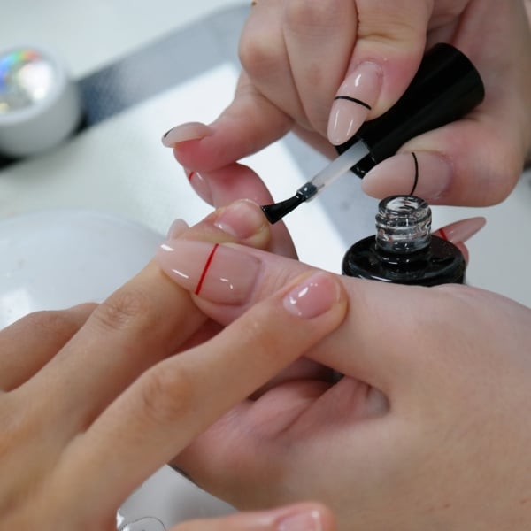 Close-up de mulher passando base fortalecedora nas unhas em casa; veste robe/camisa de tecido leve bege, segura o pincel com uma mão e aplica com o pincel na outra; foco nas mãos, fundo desfocado e ambiente aconchegante.