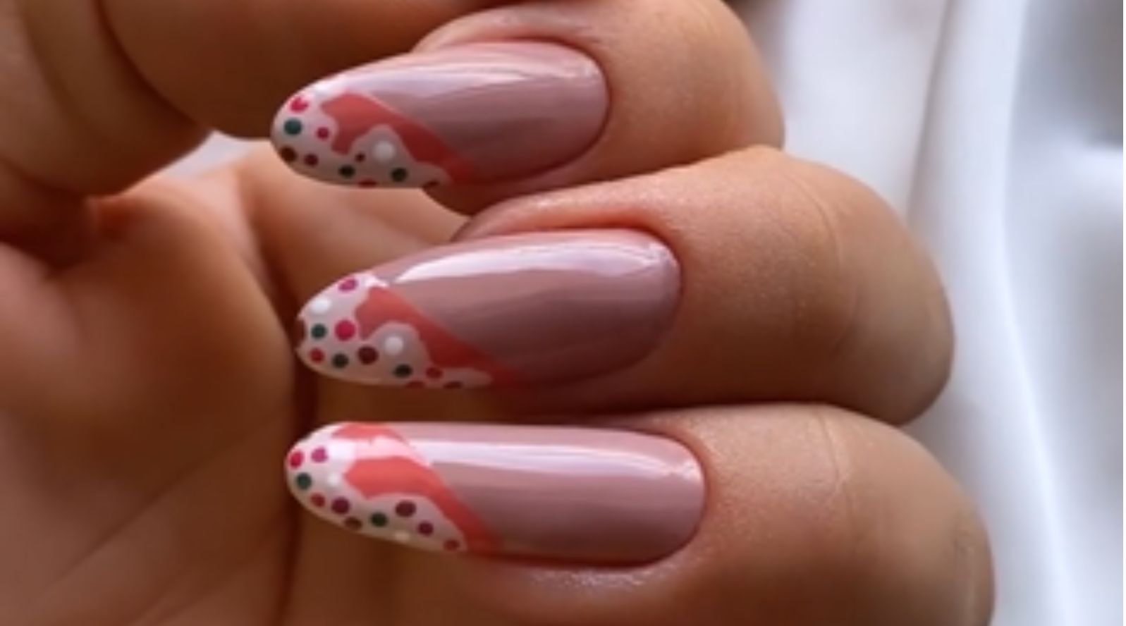 Mão com unhas ovais decoradas em base nude com pontilhismo colorido e detalhes em coral, ao lado de dois esmaltes Colorama rosa e coral.