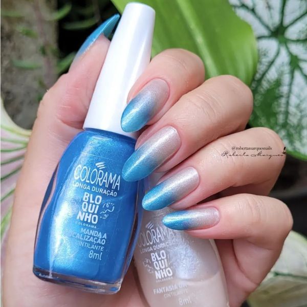 Foto de unhas feitas com esmaltes metalizados em efeito ombré, iniciando com um branco metalizado na base da unha e indo para um azul metalizado na ponta.