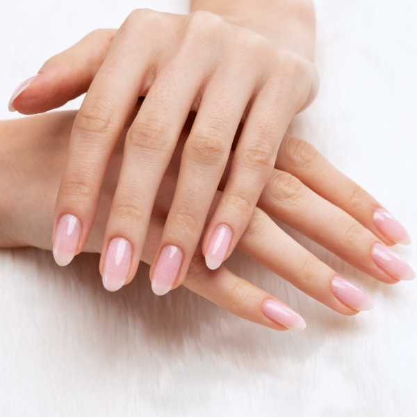 Mãos femininas sobrepostas com unhas longas em formato amendoado, esmalte rosa translúcido com acabamento perolado, remetendo à tendência soap nails delicada.