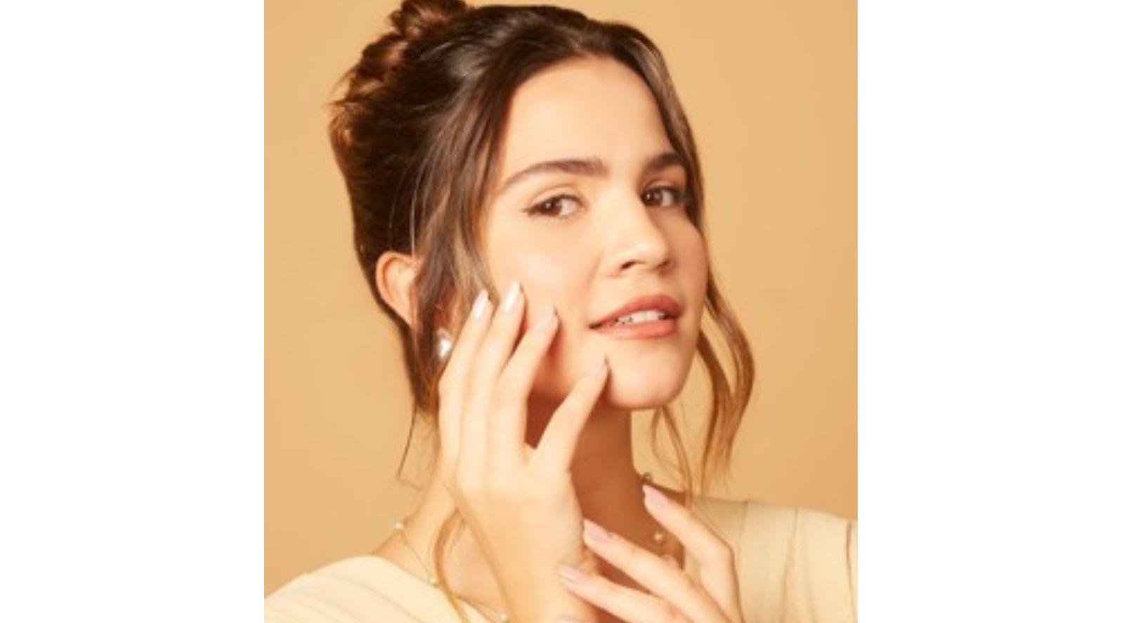 Mulher com unhas longas amendoadas em nude perolado posando com as mãos no rosto, usando joias de pérolas e blusa bege em fundo neutro.