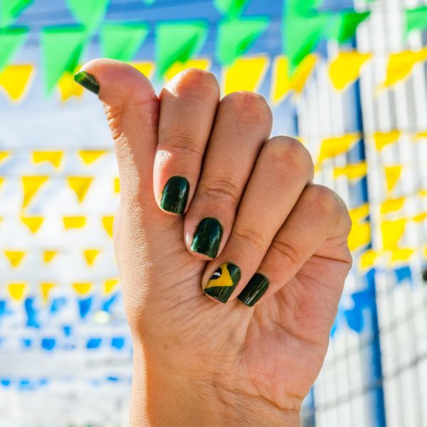 Foto de unhas feitas em esmalte verde escuro, com a filha única estampada com a bandeira do Brasil.