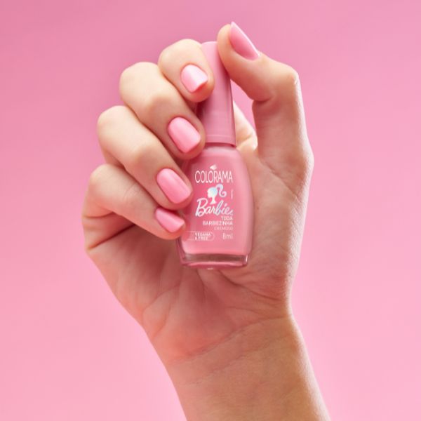 Foto de mão com o esmalte Toda Barbiezinha nas unhas rosas enquanto segura o potinho do esmalte entre os dedos.