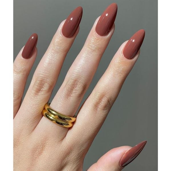 Mão com unhas simples em tom marrom e um anel dourado no dedo do meio.