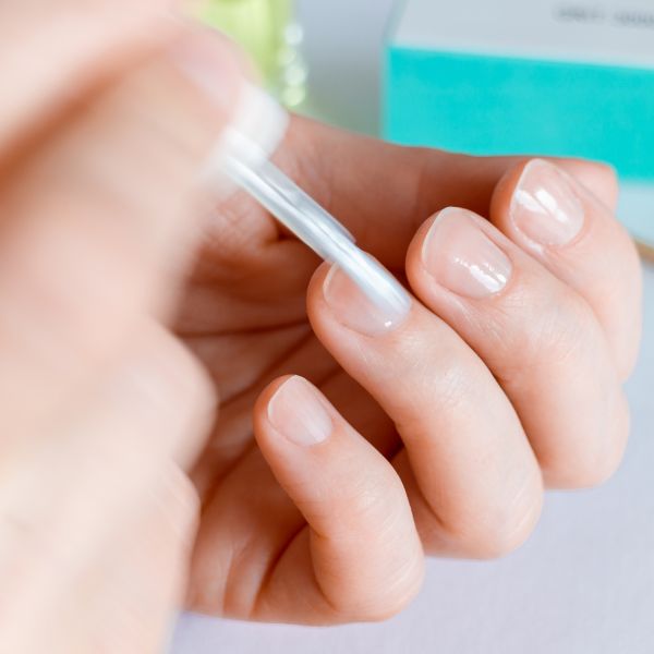 Close-up de uma mão aplicando base para unha crescer nas unhas naturais, com um pincel, sobre uma superfície clara com acessórios de manicure ao fundo.