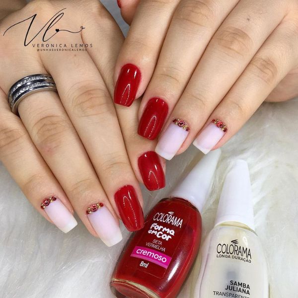 Foto de unhas feitas com esmaltação branca e vermelha cereja, com o dedo indicador e do meio em vermelho e o restante em branco com pedrarias vermelhas perto da cutícula. A foto também mostra os esmaltes Colorama utilizados no canto inferior direito.