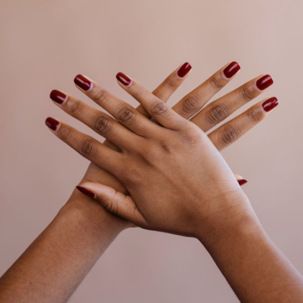 Duas mãos cruzadas com unhas longas e bem cuidadas, pintadas com esmalte vermelho vibrante, destacando a esmaltação.