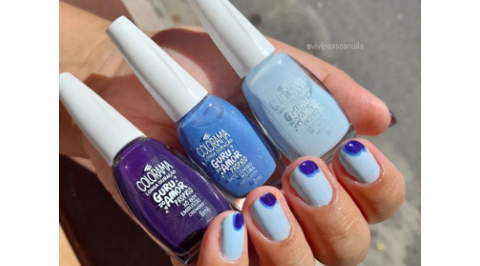 Mão com unhas curtas decoradas em azul claro com detalhes em azul royal, ao lado de três esmaltes Colorama Guru do Amor Próprio em tons de azul e roxo.
