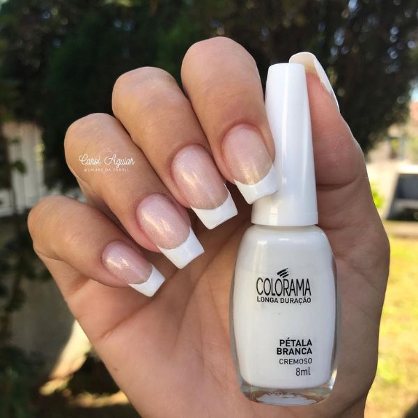 Foto de mão com unhas em formato quadrado feitas com uma base transparente com efeito shimmer e francesinhas em todos os dedos. A mão segura o frasco do esmalte “Pétala Branca” da Colorama, utilizado na esmaltação das francesinhas.
