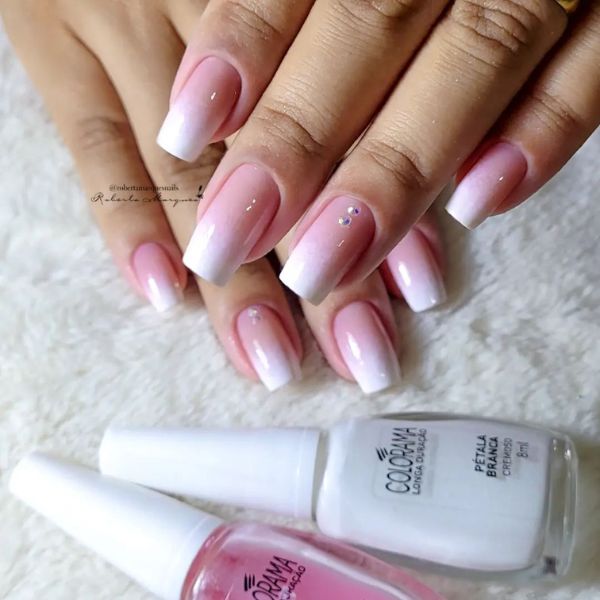 Foto de mão com unhas em formato quadrado feitas com a esmaltação estilo baby boomer, onde a cor começa rosa clara na base da unha e passa gradualmente ao branco em um estilo ombré. O dedo anelar age como filha única e, além da esmaltação baby boomer, também tem duas pedras coladas.