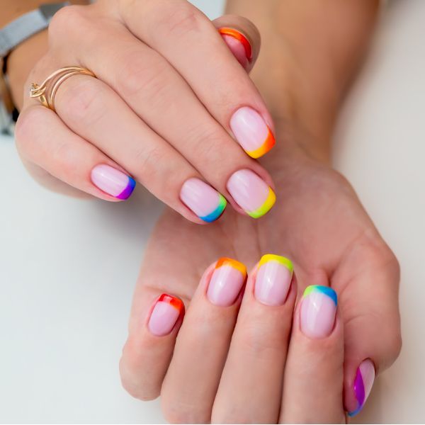 Unhas feitas com uma base rosa clara e francesinhas coloridas com uma cor em cada unha, formando um degradê das cores do arco-íris através de toda a mão.