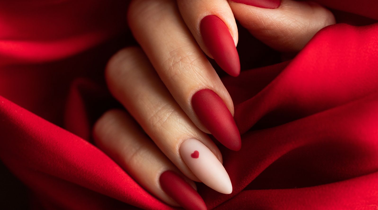 Mãos com unhas vermelhas em acabamento matte e uma unha nude decorada com coração vermelho, sobre tecido vermelho